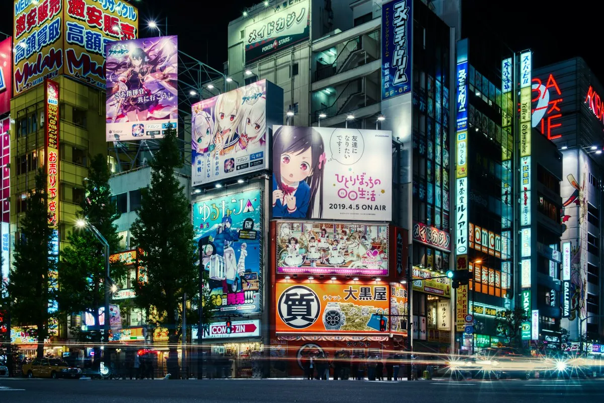 The same Chuo-dori intersection in Akihabara after dark, with lit billboards and light trails from passing cars