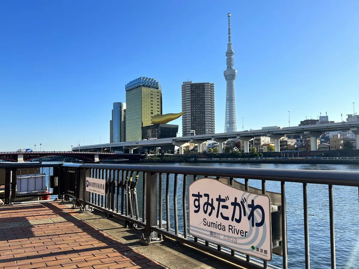 A Sumida River sign on the riverside walkway in Asakusa, with Tokyo Skytree across the water under a clear sky