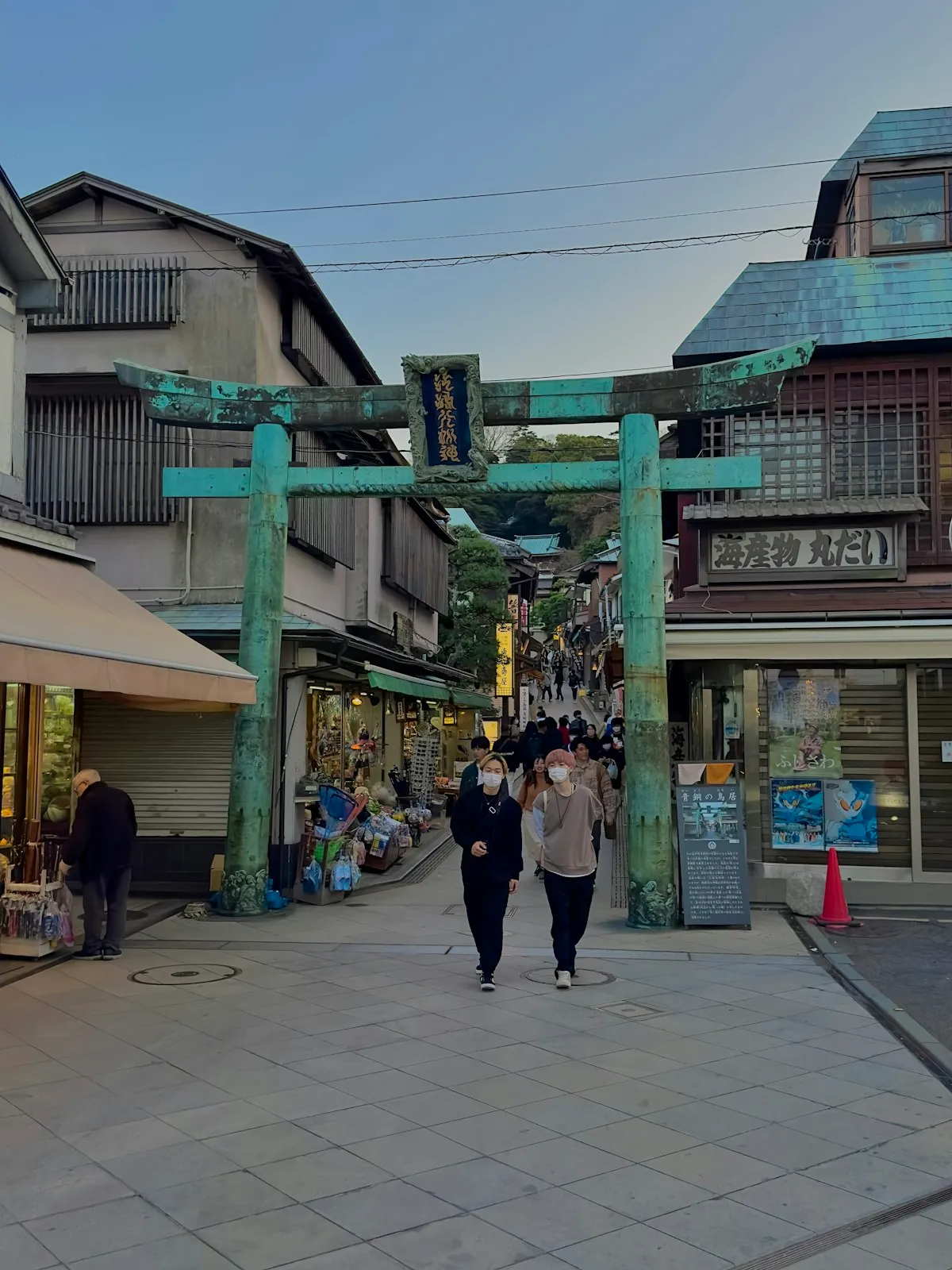 The weathered bronze torii at the foot of Enoshima's shopping street, with the lane climbing uphill behind it