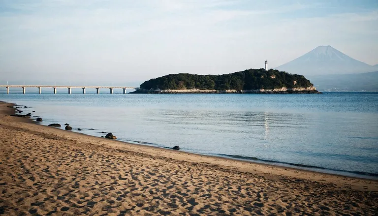 Enoshima island seen across the water from the Shonan coast, with a small bridge leading to it and a hazy Mount Fuji on the horizon