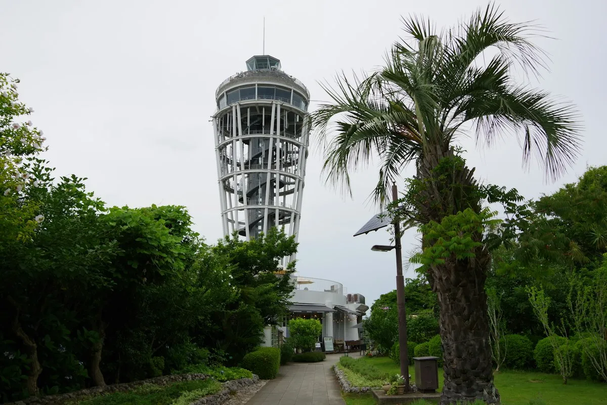 The Enoshima Sea Candle lighthouse tower beyond a garden path and a palm tree on an overcast day