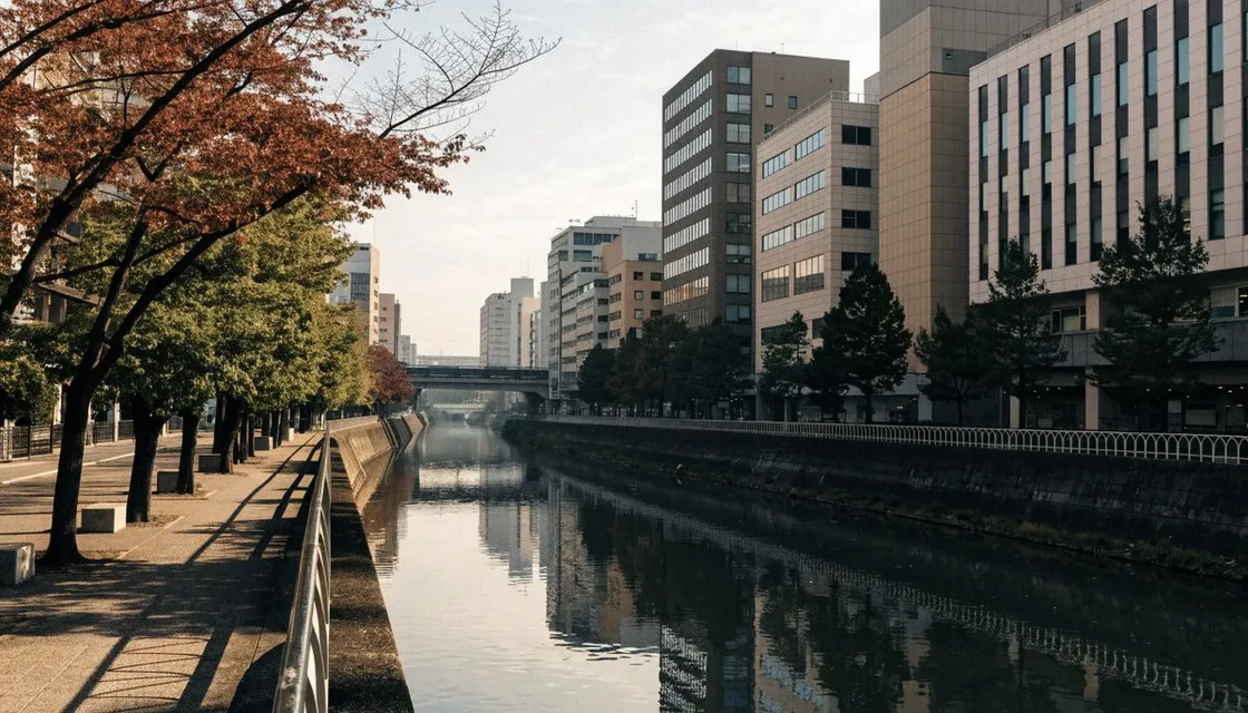 朝のやわらかな光の中、木々と低い柵に沿って流れる都市の川。奥に中層のオフィスビルが並ぶ