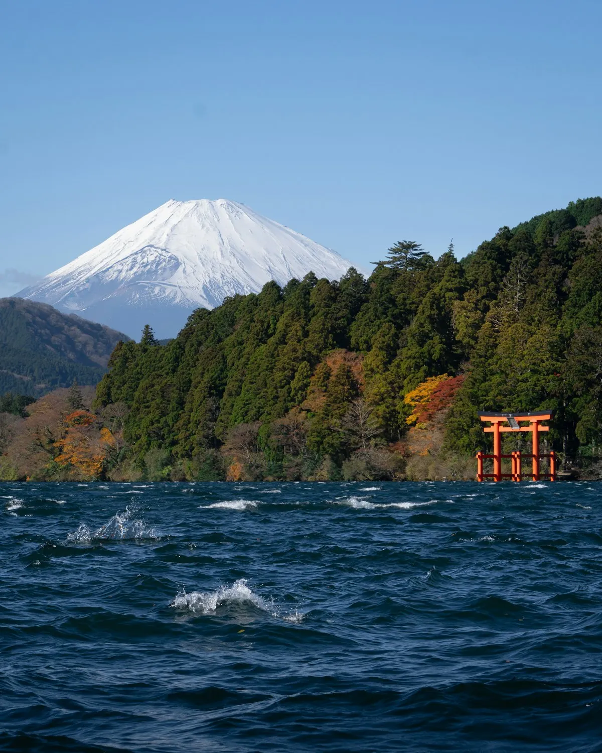 芦ノ湖の森の向こうにそびえる雪化粧の富士山と、水辺に立つ赤い鳥居