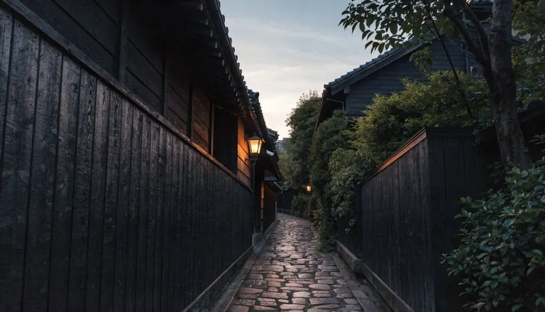 夕暮れの神楽坂の細い石畳の路地。黒板塀にはさまれた坂道に、灯りがひとつともる風景