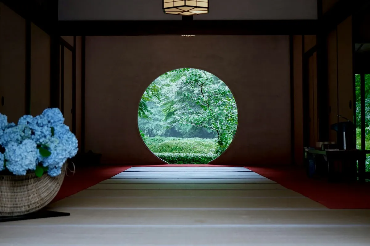 A round window at a Kamakura temple looking out onto a green garden, with a basket of blue hydrangeas in the room