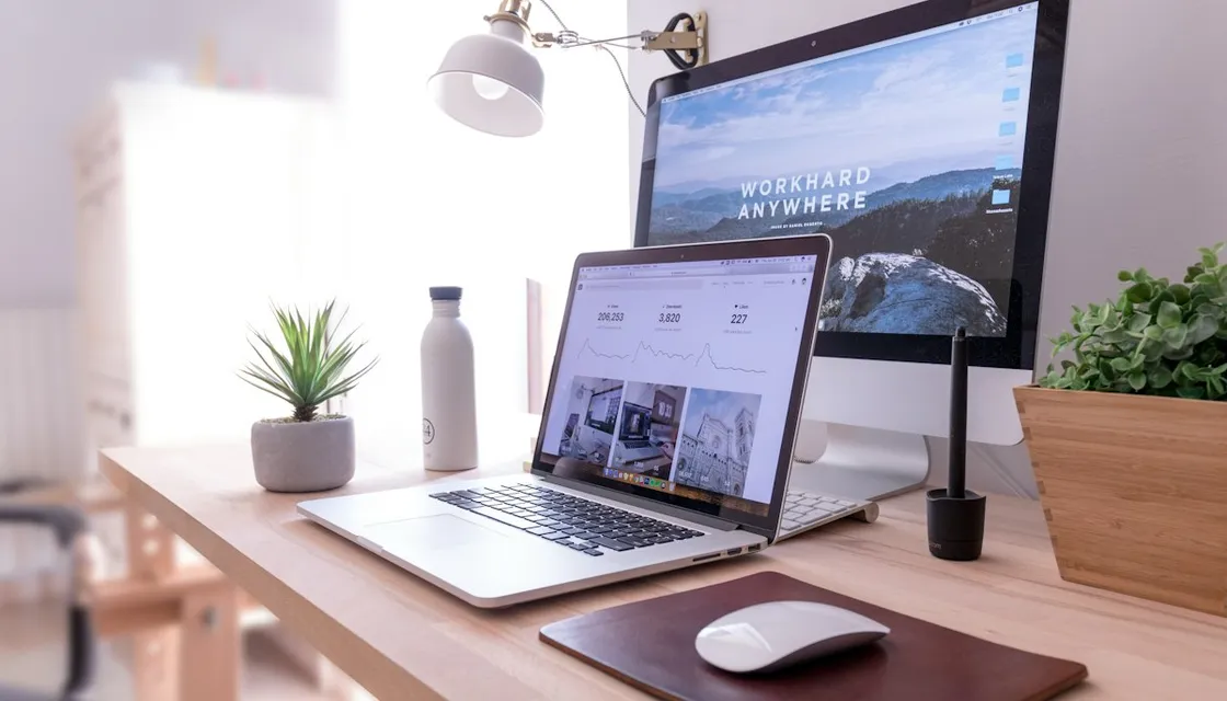 A laptop and external monitor on a wooden desk with small plants