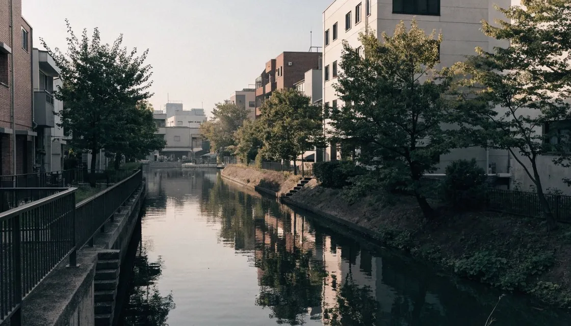 低層の建物にはさまれた細い川沿いの遊歩道。手すりが続き、水面が空を映している平日の静かな風景