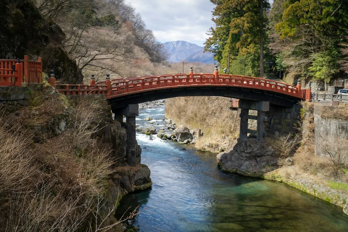 The red-lacquered Shinkyo Bridge over a clear river in Nikko, with mountains in the distance
