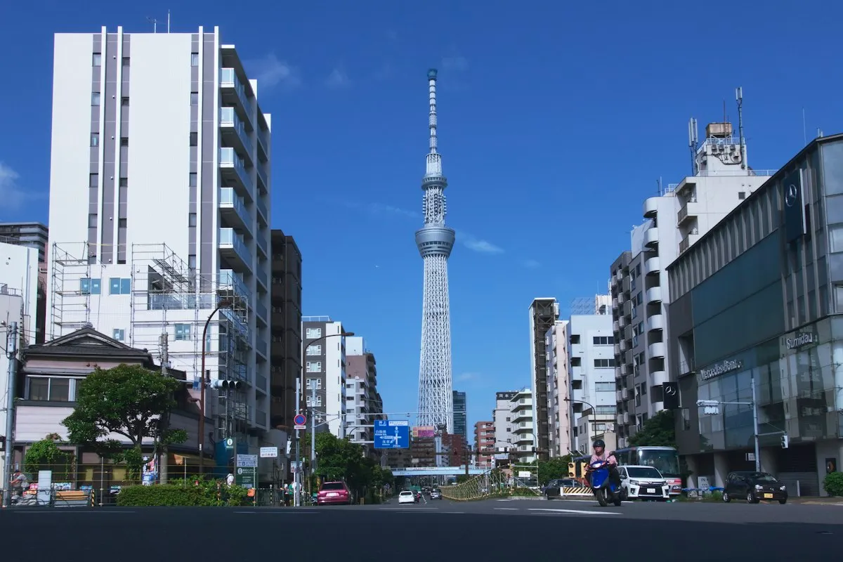 A wide street lined with mid-rise buildings under a blue sky, with Tokyo Skytree rising at the end