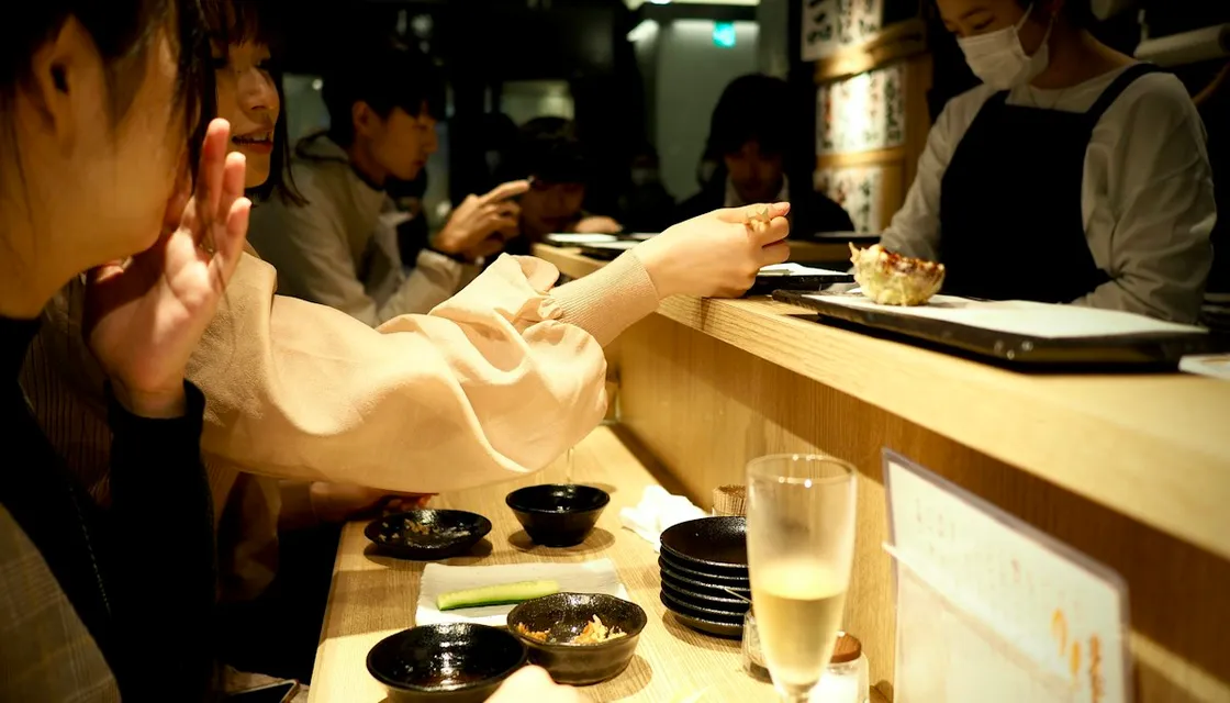 Customers eating and drinking at a busy wooden counter in a Tokyo bar